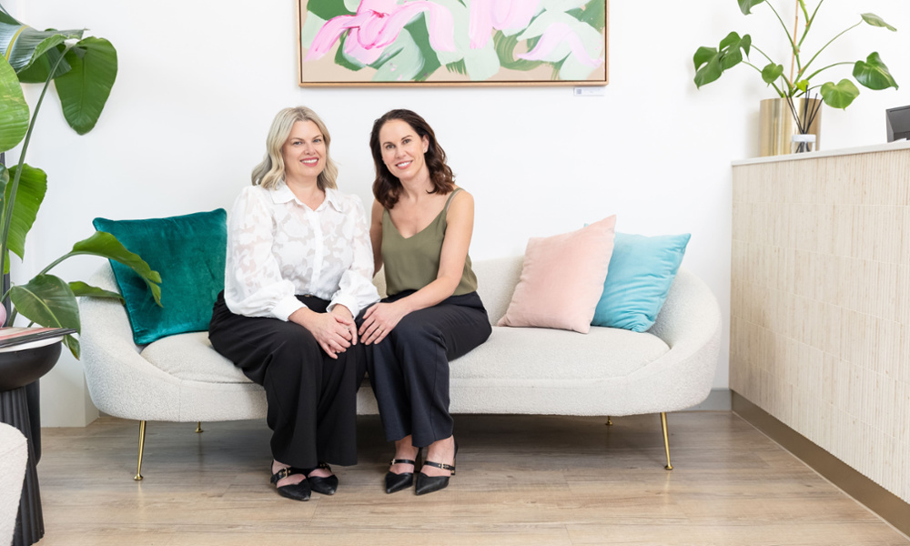Co-founders Dr Meredith Metin and Rachael Jones seated together on a modern sofa in a bright, stylish dental practice waiting area.